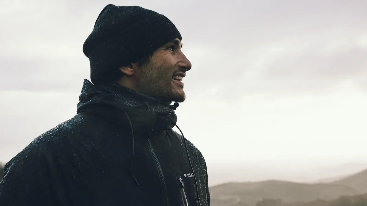 randonneur souriant en bonnet et veste noire face aux montagnes sous un ciel voilé
